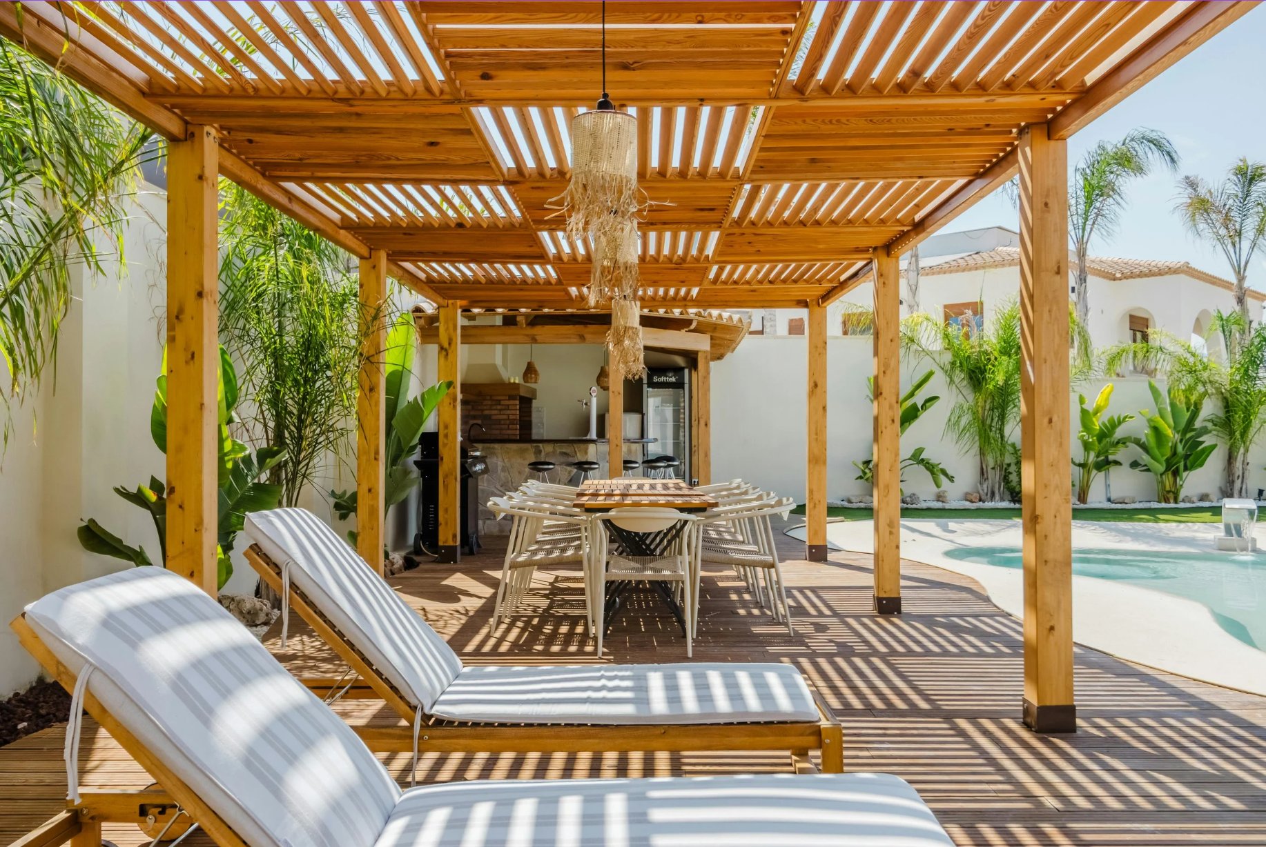 A pergola-covered terrace with timber slats, a long table, and a pool