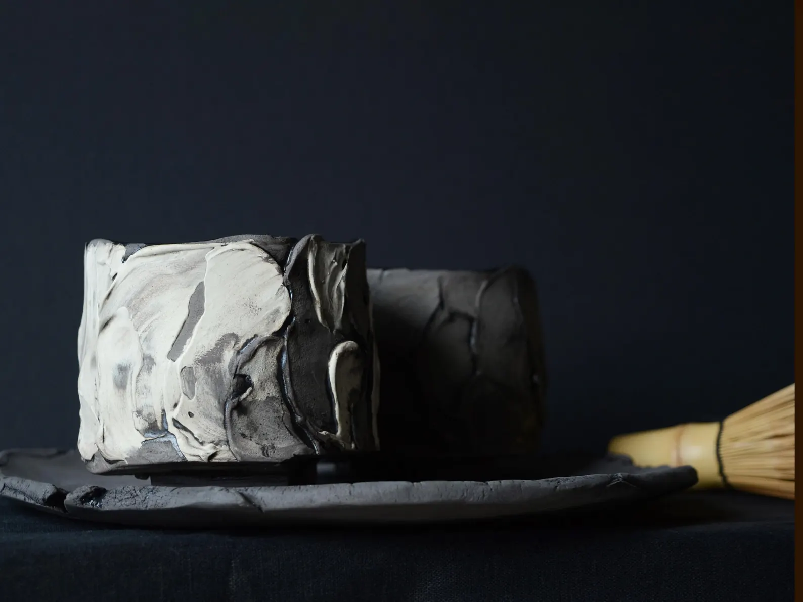 A black-glazed stoneware teabowl with cream marbling on a clay tray, with a bamboo whisk